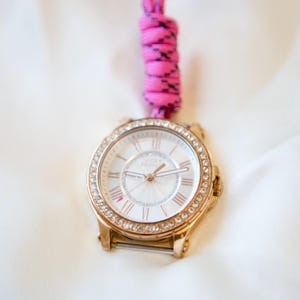 Peut inclure: Une montre-bracelet dorée avec un cadran rond, une lunette ornée de cristaux transparents et un cadran blanc avec des chiffres romains et le texte Juicy Couture. Un cordon tressé rose vif et noir est attaché au sommet du boîtier de la montre.