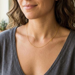 Puede incluir: Un delicado collar dorado con un colgante con la letra "A". El colgante está situado a un lado del collar. El collar lo lleva una persona con una camiseta gris.