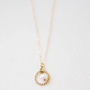 Pode incluir: Um delicado colar de ouro com um pequeno pingente de pérola branca e redonda. O pingente é circundado por um anel de ouro torcido, suspenso de uma fina corrente de ouro. O colar é exibido em um fundo branco macio.