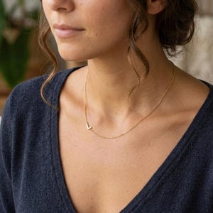 May include: A delicate gold-colored necklace featuring the letter "L" as a pendant. The necklace is worn on a person's neck, with the pendant positioned to the side. The chain is thin and elegant, creating a minimalist and stylish look.