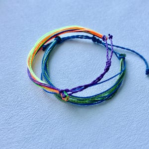 May include: A set of colorful friendship bracelets. The bracelets feature a combination of vibrant colors, including blue, green, purple, orange, and yellow. The bracelets are made of woven threads and are tied with knots.