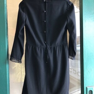 May include: A black dress with long sleeves and a sheer lace trim at the neckline and hem. The dress has a button closure down the back.