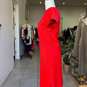 May include: A vibrant red dress displayed on a mannequin. The dress features a fitted bodice with short sleeves and a knee-length skirt with a slit. The dress is made of a smooth fabric and is a solid, bright red color. The dress is displayed in a retail setting.