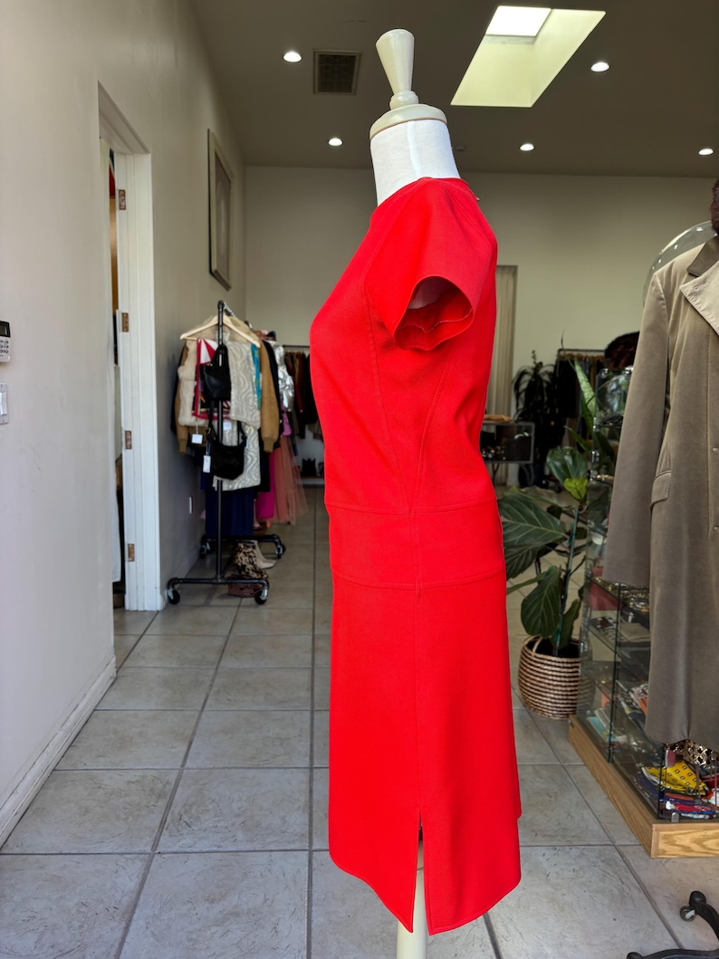 May include: A vibrant red dress displayed on a mannequin. The dress features a fitted bodice with short sleeves and a knee-length skirt with a slit. The dress is made of a smooth fabric and is a solid, bright red color. The dress is displayed in a retail setting.
