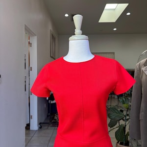 May include: A vibrant red dress with short sleeves and a round neckline. The dress is displayed on a mannequin in a boutique setting. The dress has a fitted bodice and a skirt that appears to be knee-length.