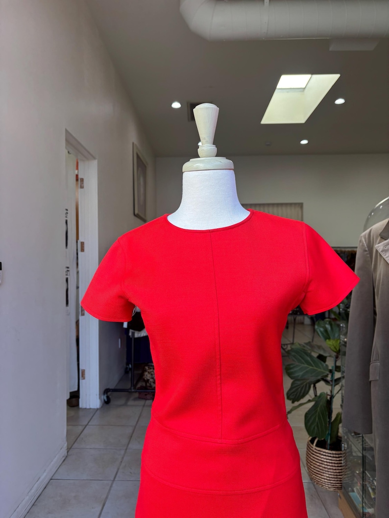 May include: A vibrant red dress with short sleeves and a round neckline. The dress is displayed on a mannequin in a boutique setting. The dress has a fitted bodice and a skirt that appears to be knee-length.