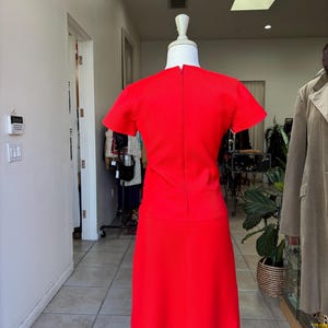 May include: A vibrant red dress with short sleeves and a knee-length skirt. The dress is displayed on a mannequin in a well-lit boutique setting. The dress has a simple, classic design, perfect for a vintage or retro aesthetic.