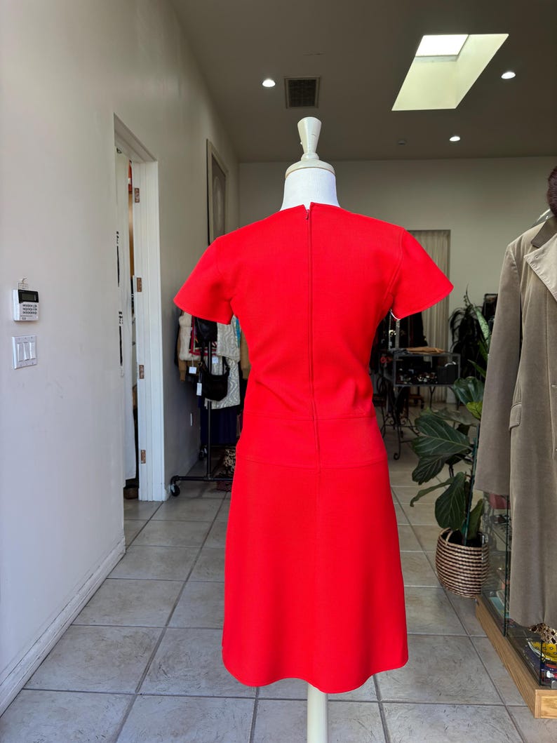 May include: A vibrant red dress with short sleeves and a knee-length skirt. The dress is displayed on a mannequin in a well-lit boutique setting. The dress has a simple, classic design, perfect for a vintage or retro aesthetic.