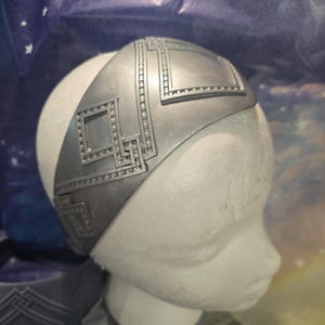 Puede incluir: Una diadema plateada con un diseño Art Déco. La diadema está hecha de un material rígido y presenta patrones geométricos y formas rectangulares. La diadema se muestra en una cabeza de maniquí blanca.