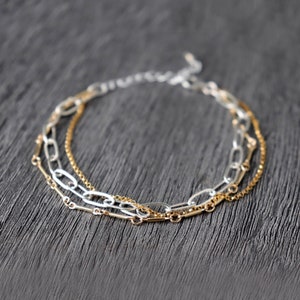 May include: A layered chain bracelet featuring three different chain styles. The bracelet is made of silver and gold metal.