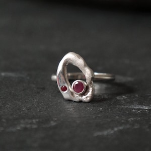May include: A silver ring with an abstract design, featuring two round, red gemstones. The ring has a textured, organic shape with a smooth band. The gemstones are set in the silver, creating a striking contrast.