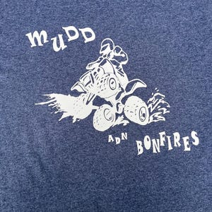 May include: A navy blue t-shirt with a white graphic of a person riding an ATV, with the words "mudd adn bonfires" in a stylized font. The shirt is made of a soft material.