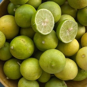 May include: A wooden bowl filled with fresh limes, some whole and some halved. The limes range in color from bright green to yellow-green, with a few showing a yellow hue. The cut limes reveal juicy, pale green flesh.