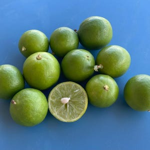 May include: A collection of fresh, green limes, some whole and some halved, arranged on a bright blue surface. The limes are vibrant green with a smooth texture, showcasing their natural freshness. One lime is cut in half, revealing the juicy interior.