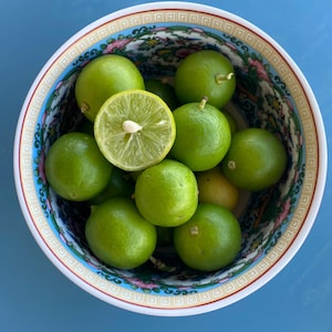 May include: A decorative bowl filled with fresh green limes, one cut in half. The bowl has a white rim with a blue, red, and gold patterned design. The limes are a vibrant green color.