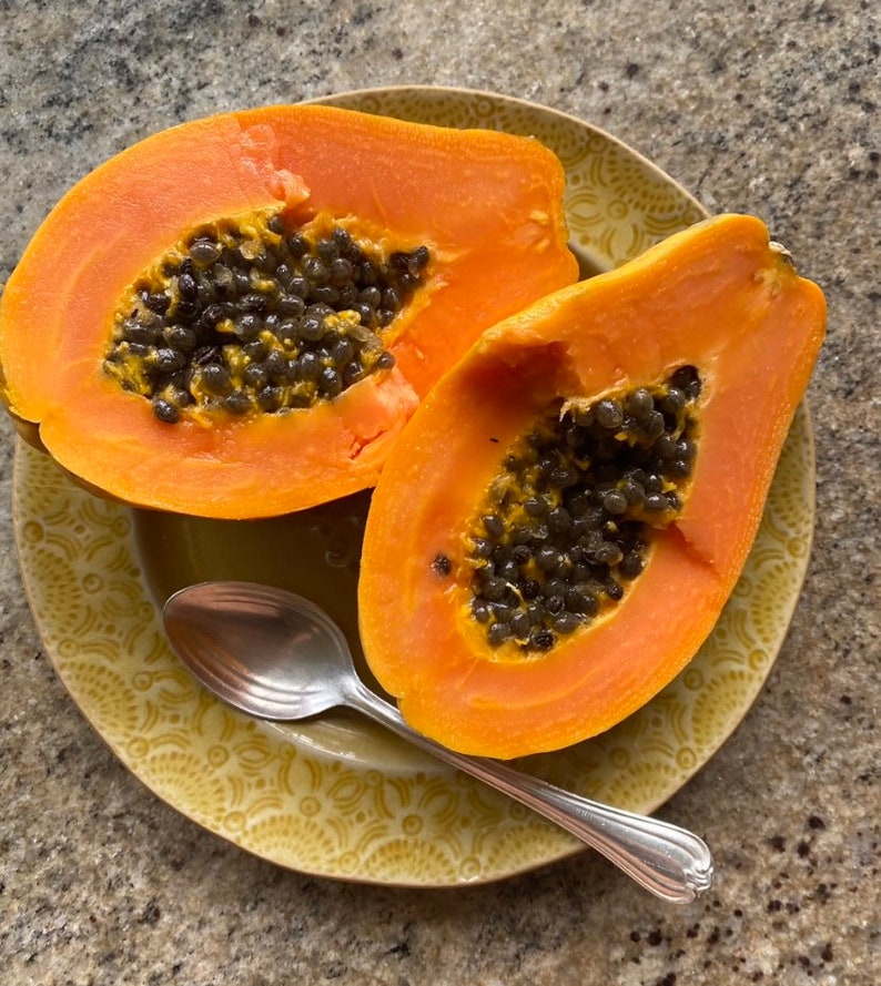 May include: Two halves of a ripe papaya on a yellow patterned plate with a silver spoon. The papaya is orange with black seeds.