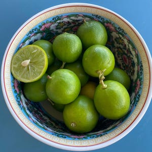 May include: A decorative bowl filled with bright green limes, one cut in half. The bowl has a detailed floral pattern with pink, blue, and gold accents. The background is a solid blue surface.