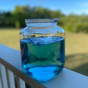 Puede incluir: Una jarra de vidrio transparente llena de un l&iacute;quido azul brillante. La jarra tiene una superficie texturizada y est&aacute; sentada sobre una barandilla de madera blanca.