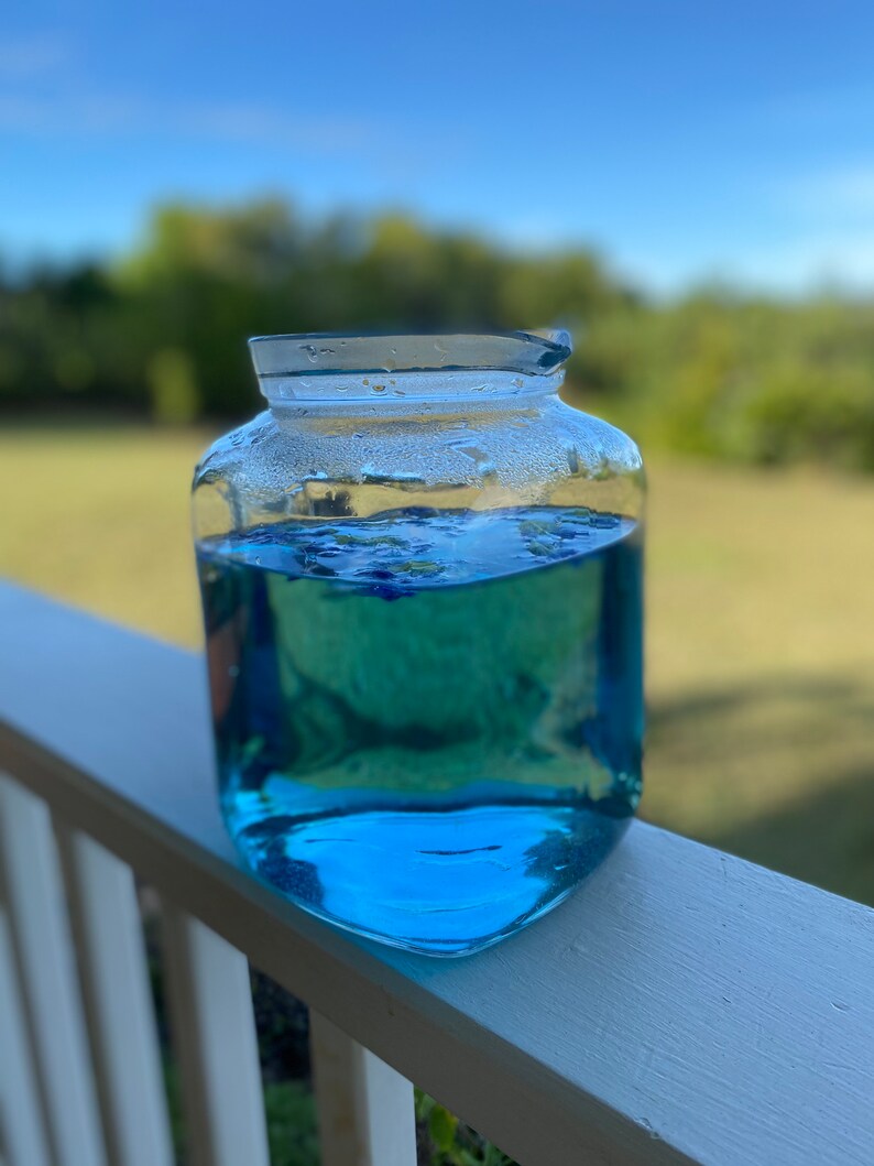 Puede incluir: Una jarra de vidrio transparente llena de un l&iacute;quido azul brillante. La jarra tiene una superficie texturizada y est&aacute; sentada sobre una barandilla de madera blanca.