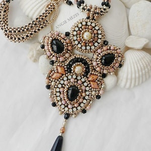 Op de afbeelding: Een kralenketting met een grote, ingewikkelde hanger bestaande uit zwarte, perzikkleurige en metallic gouden kralen met centrale zwarte druppelvormige en ovale stenen. De tekst ANGIE MEZES is zichtbaar op de hanger. Inclusief bijpassende kralenketting.