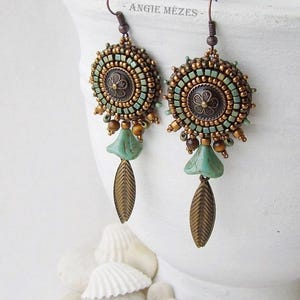May include: A pair of handcrafted earrings with a bohemian design. The earrings feature a circular beaded design in shades of teal, bronze, and brown, with a central floral element. Dangling from the earrings are teal teardrop-shaped beads and bronze leaf charms.