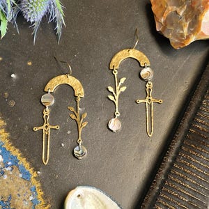 May include: A pair of gold-colored earrings with a unique design. Each earring features a curved top, a small gemstone, a sword pendant, and a delicate leaf-like element. The earrings are displayed on a dark surface with decorative elements.