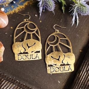 May include: Gold-colored earrings with a sun and moon design, featuring the word "DOULA" at the bottom. The earrings are displayed on a dark brown surface with decorative elements, including flowers and stones.