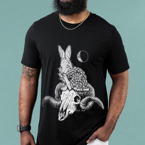 May include: Black t-shirt with a white graphic of a rabbit sitting on a skull with horns. The graphic also includes a crescent moon.