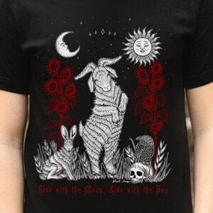 May include: Black t-shirt with a white graphic design featuring a goat, a rabbit, a raven, a skull, a crescent moon, and a sun with a face. The text "Rise with the Moon, Rise with the Sun" is printed in red at the bottom of the design.