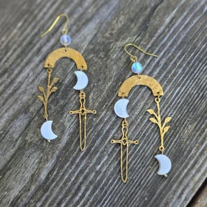 May include: A pair of gold-colored earrings with a celestial theme. Each earring features a hammered gold arch, a crescent moon, a sword pendant, and a sprig of leaves. The earrings are approximately 3 inches long.