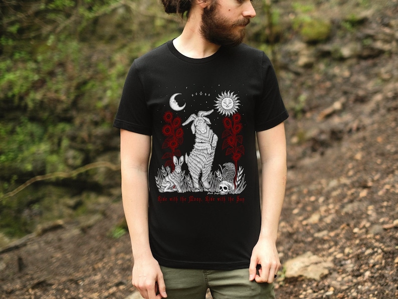 May include: Black t-shirt with a white graphic design featuring a goat, a rabbit, and a skull. The graphic also includes a crescent moon, a sun, and the text "Rise with the Moon, Rise with the Sun".