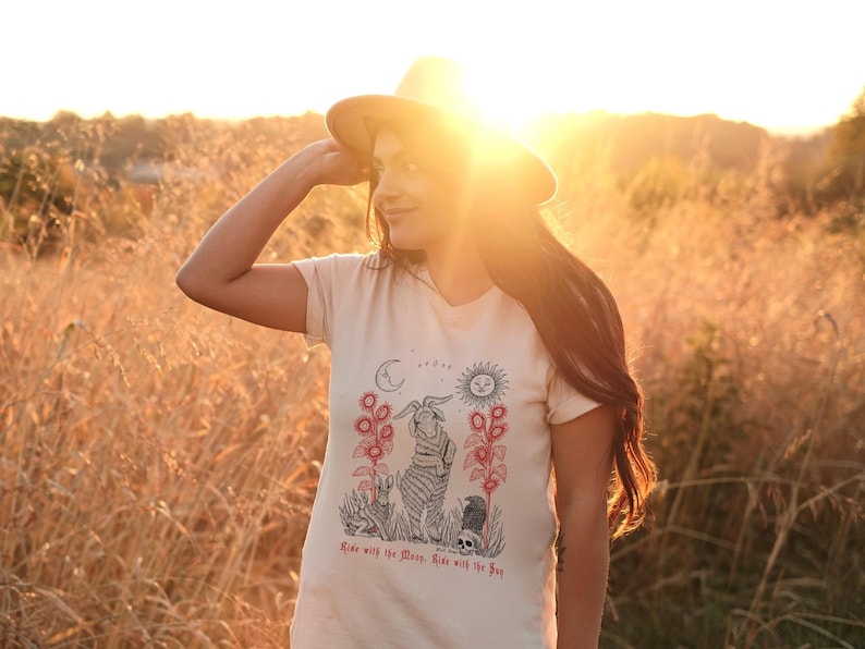May include: A woman wearing a beige t-shirt with a black and white illustration of a creature with a moon and sun above it. The text on the shirt reads "Rise with the Moon. Rise with the Sun."