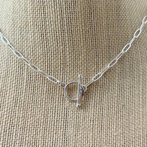 May include: A silver necklace with an oval link chain and a toggle clasp. The necklace is displayed on a neutral-colored linen surface. The toggle clasp is a circular ring with a straight bar.