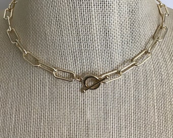 May include: Gold-colored chain necklace with large rectangular links. The necklace features a toggle clasp closure. The jewelry is displayed on a neutral-colored mannequin.