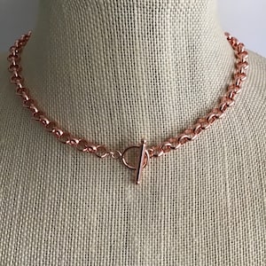 May include: Rose gold chain necklace with a toggle clasp. The necklace features a series of rounded links, creating a chunky, yet delicate appearance. The toggle clasp is a simple bar and ring design. The necklace is displayed on a neutral-toned mannequin.