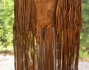 May include: A brown suede vest with long fringe detailing. The vest is hanging on a wooden hanger. The fringe hangs down the front and back of the vest, and the vest has a tie closure at the waist.