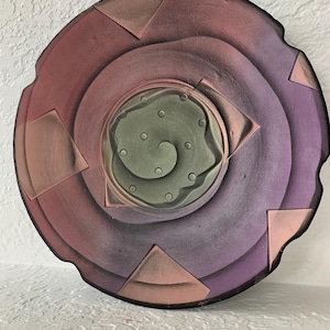 May include: A circular ceramic wall plate with a swirling, abstract design in shades of pink, purple, and green. The center of the plate features a raised, textured design with a spiral pattern.