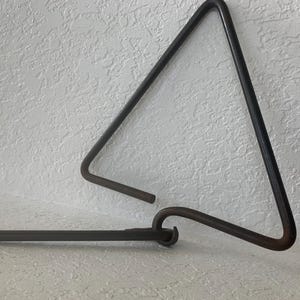 May include: A dark gray metal triangle musical instrument with a smooth, rounded finish. The instrument is shaped as an open-ended triangle with one corner slightly curved, and it includes a matching straight metal striker rod.