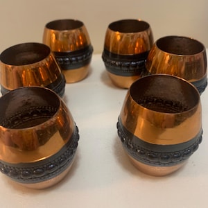 May include: A set of six small, barrel-shaped cups featuring a polished copper finish on the top and bottom sections, separated by a central dark, textured band with a raised, beaded pattern encircling the middle of each individual vessel.