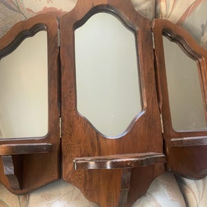 May include: A vintage wooden vanity mirror with three hinged panels. The mirror has a dark brown finish and decorative, scalloped edges. Each panel has a mirror, and the center panel has a small shelf. The side panels also have small shelves.