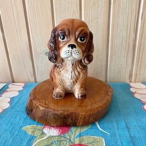 Op de afbeelding: Een bruin en wit keramiek beeldje van een hond die op een houten schijf zit. De hond heeft bruin haar, een witte borst en zwarte ogen.