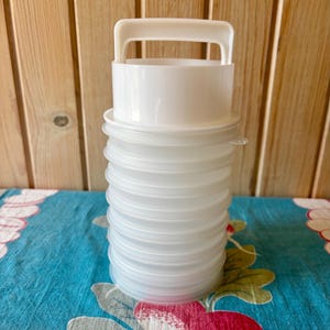 May include: A white plastic stackable container set featuring a cylindrical design with a top handle. The unit consists of multiple translucent, circular, snap-on lids or shallow trays stacked vertically to form a single storage tower structure.