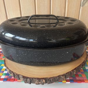 May include: A black speckled enamel roasting pan with a lid and handle. The oval-shaped pan sits on a wooden slice and a colorful woven mat. The lid has several small vent holes.