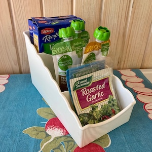 Puede incluir: Organizador de almacenamiento de plástico blanco de cuatro niveles. El compartimento superior contiene una caja de bolsitas de té Lipton Recipe. Los otros compartimentos contienen cuatro pequeñas bolsas de comida para bebés de color verde y blanco y un pequeño paquete de condimento Good Seasons Roasted Garlic.
