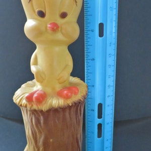 May include: A yellow rubber Tweety Bird toy sitting on a brown tree stump. Tweety is wearing a red and black hat and has a red beak.