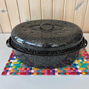 May include: Black speckled enamel roasting pan with a lid, featuring a handle on top and side handles. The pan sits on a colorful woven mat. The pan is made in the USA.