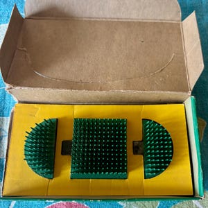 May include: A vintage floral arranging kit with three green pin holders in a yellow and brown cardboard box. The pin holders are a square and two half-circle shapes, all with numerous upward-facing pins.