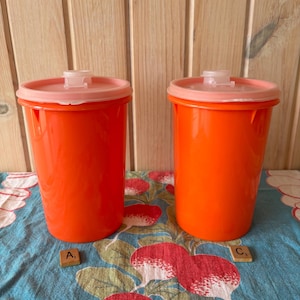 May include: Two vintage orange plastic containers with pink lids. The containers are cylindrical and appear to be in good condition. The lids have a small tab for opening. The containers are on a blue floral tablecloth.