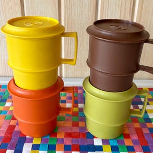 May include: Two sets of vintage plastic mugs in bright colors. One set features a yellow mug stacked on an orange mug, and the other has a brown mug on a green mug. Each mug has a handle and a lid.