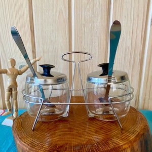 Peut inclure: Un ensemble de deux pots à condiments en verre avec des couvercles et des cuillères en argent, sur un support en argent. Les pots sont transparents et les cuillères sont attachées aux couvercles.